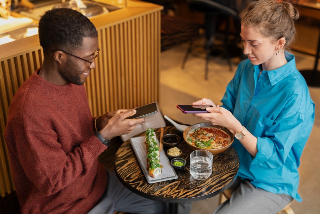 Um homem e uma mulher sentados em frente a uma mesa fotografam seus pratos de comida.